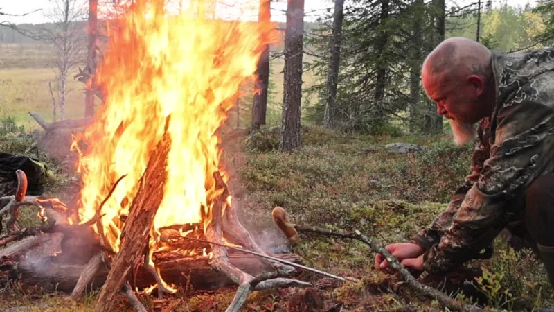 Partasuu metsästäjä kääntää makkaraa nuotion loimussa